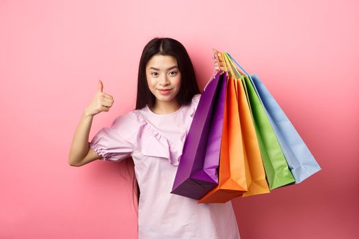 Shopping. Stylish girl showing colorful paper bags and thumbs-up, recommend store discounts, standing against pink background.