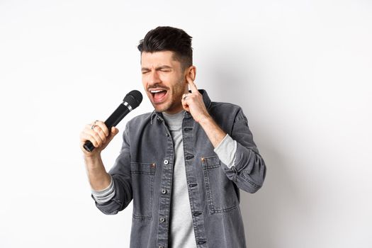 Handsome singer trying to reach high note, singing in microphone and shut one ear, standing on white background.