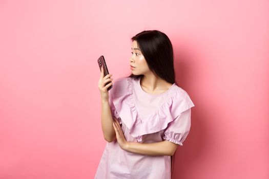 Asian girl look surprised at smartphone screen, reading message, standing in dress against pink background.