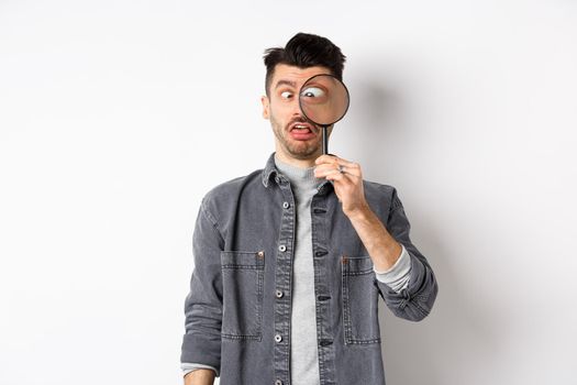 Funny young man squinting, showing faces with magnifying glass, standing on white background.