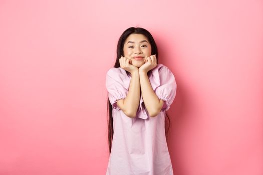 Cute and happy asian woman waiting for something with excitement, lean face on hands and watching lovely scene with admiration, standing against pink background.