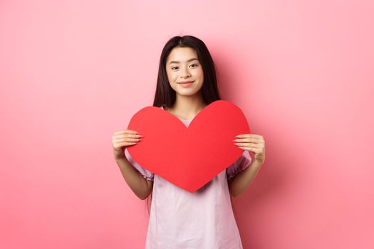Valentines day concept. Cute teenage asian girl showing big red heart card, falling in love, going on romantic date in dress, smiling tender at camera, pink background.