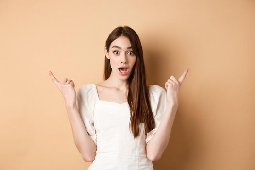 Surprised young girl pointing sideways at left and right, gasping and saying wow amazed, showing two cool choices, beige background.