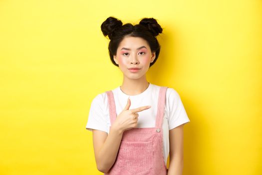 Stylish asian teen girl with makeup and summer clothes, pointing finger right and look serious, standing against yellow background.