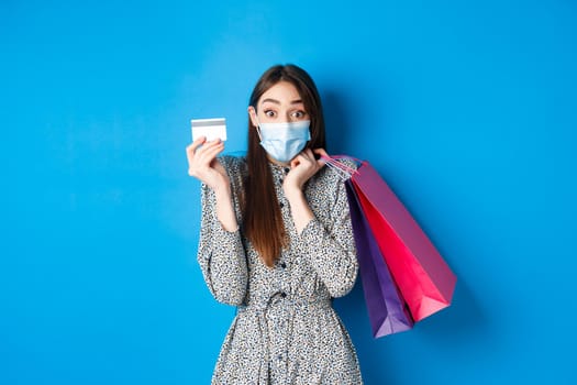 Covid-19, pandemic and lifestyle concept. Excited woman wearing medical mask while shopping, showing plastic credit card and holding bags, blue background.