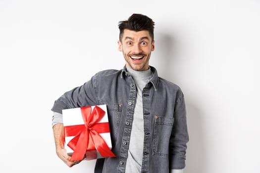 Happy Valentines day. Surprised and happy guy holding gift box and look at camera, smiling amazed, celebrating holiday, bring present on romantic date, white background.