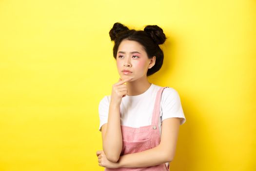 Thoughtful serious girl with beauty makeup, looking left and thinking with suspicious face, have doubts, standing on yellow background.