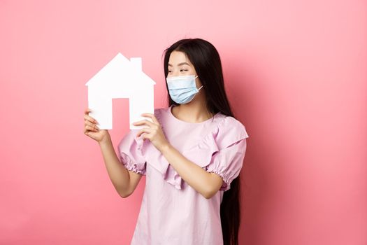 Real estate and pandemic concept. Excited asian girl in medical mask looking pleased at paper house cutout, buying property, standing on pink background.