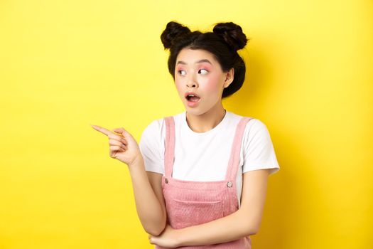 Intrigued asian beauty girl with makeup, looking curious at logo, pointing finger aside, standing in summer clothes against yellow background.