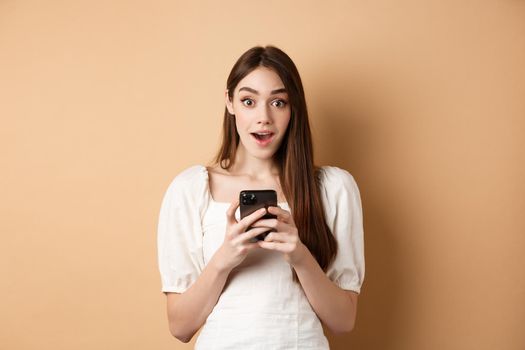 Exciting news on phone. Amazed young woman say wow after reading online announcement on smartphone, standing on beige background.