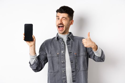 Handsome guy winking and showing empty cell phone screen with thumb up, recommending app or online deal, standing against white background.