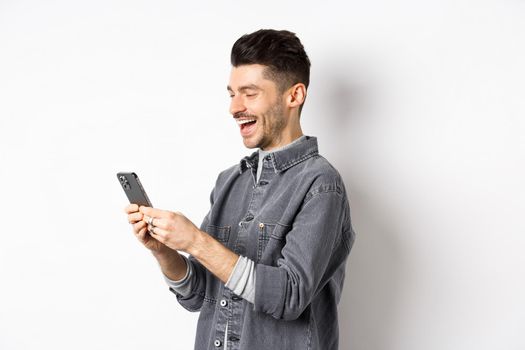 Profile of handsome man chatting on phone, laughing and reading smartphone screen, standing on white background.