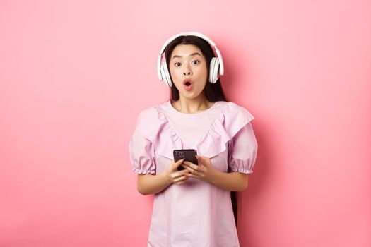 Excited asian woman in wireless headphones say wow, holding mobile phone and looking amused at camera, standing against pink background.