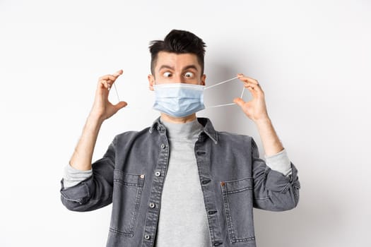 Health, covid and pandemic concept. Funny man squinting eyes at medical mask, playing with it, standing on white background.