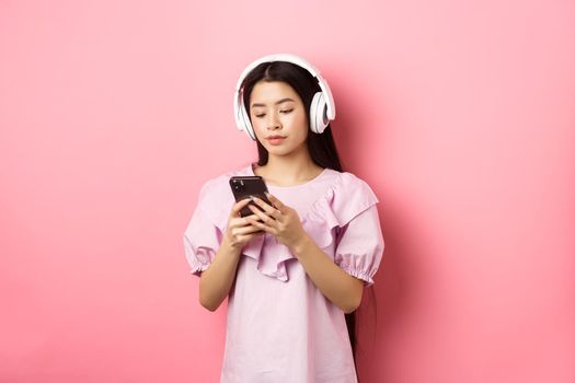 Young girl listening music in headphones and chatting on mobile phone, standing in dress against pink background.