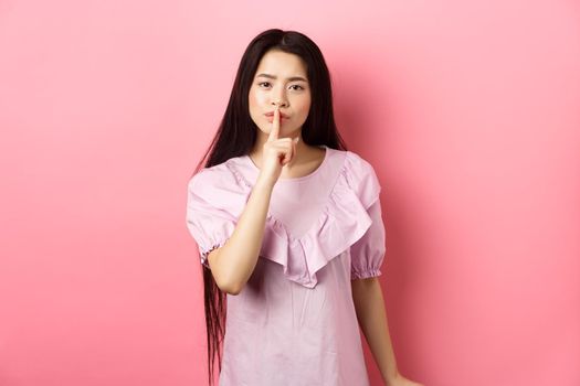 Cute asian girl tell to be quiet, scolding loud person, showing shush sign with finger pressed to lips, standing in dress on pink background.