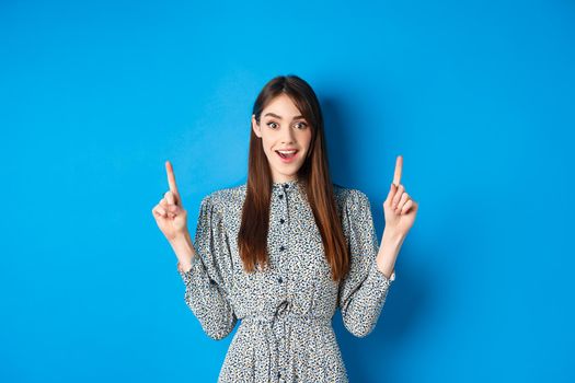 Excited and happy young woman in dress checking out special offer, pointing fingers up and smiling, standing against blue background.