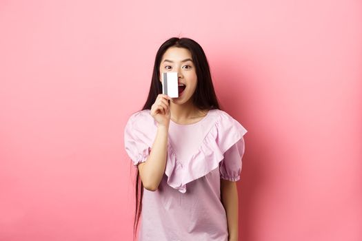 Excited asian girl showing plastic credit card and smiling, going on shopping, standing against pink background.