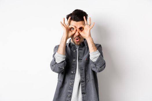 Funny guy look through hand binoculars, checking out awesome promo, say wow and stare at camera excited, standing on white background.