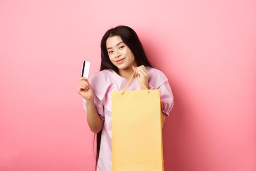 Shopping. Stylish asian girl showing shop bag and plastic credit card, standing on pink background.