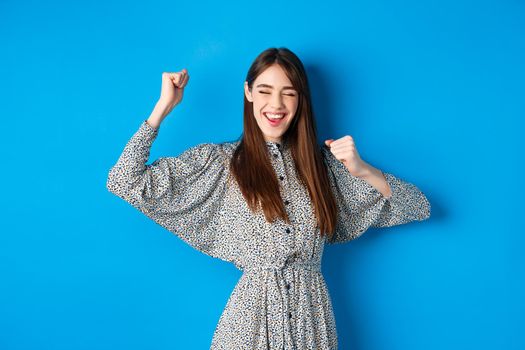 Excited smiling girl showing tongue and dancing with raised hands, celebrating victory or achievement, winning prize, standing happy on blue background.