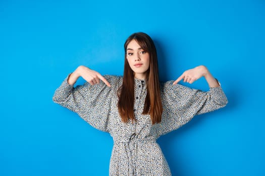 Confident pretty girl in dress pointing fingers at center logo, showing banner and looking self-assured, standing on blue background.