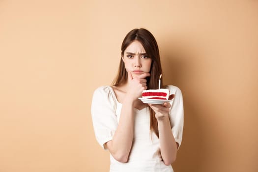 Thoughtful birthday girl frowning and thinking of wish, holding cake with candle, standing on beige background.