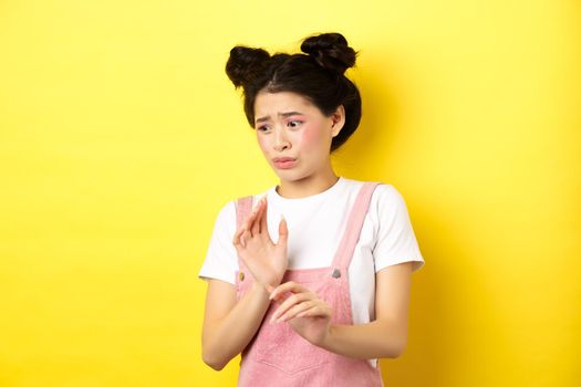 Disgusted asian girl raising hands up defensive, block something disgusting, turn away from aversion and reluctance, standing on yellow background.