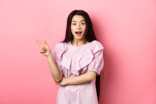 Excited beautiful asian teen girl pointing finger left, open mouth amazed and looking at camera, showing cool promo deal, pink background.