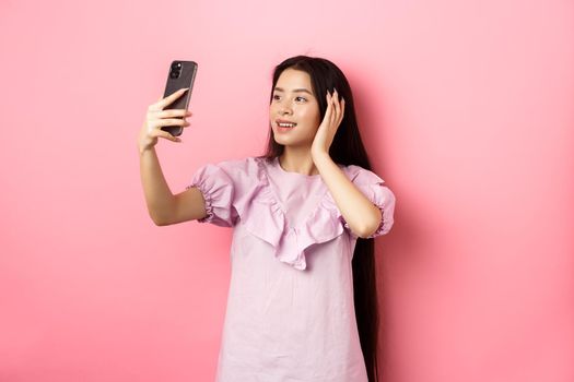 Stylish asian girl blogger taking selfie on mobile phone, posing for smartphone photo, standing in dress against pink background.