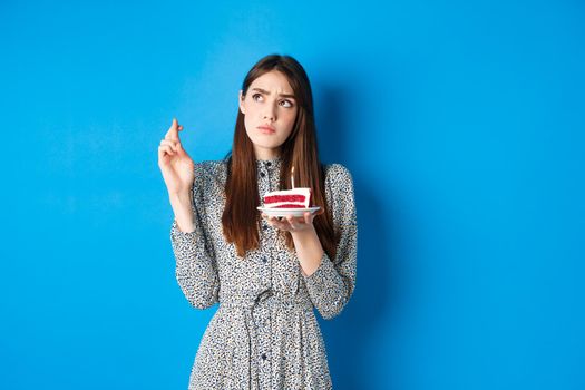 Thoughtful birthday girl making wish, cross fingers good luck, holding cake and looking at upper left corner, blue background.