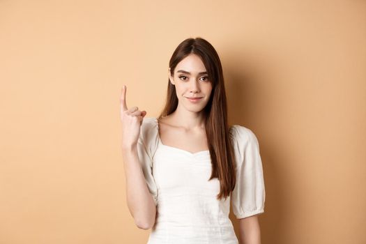 Attractive young woman show finger number one, smiling and looking confident, standing on beige background.