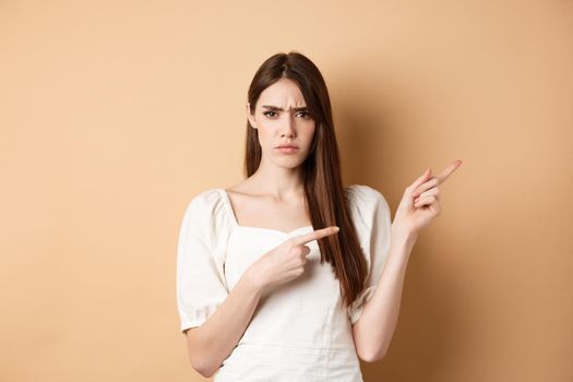 Angry young woman frowning, pointing fingers right and complaining, disappointed with logo, standing upset on beige background.