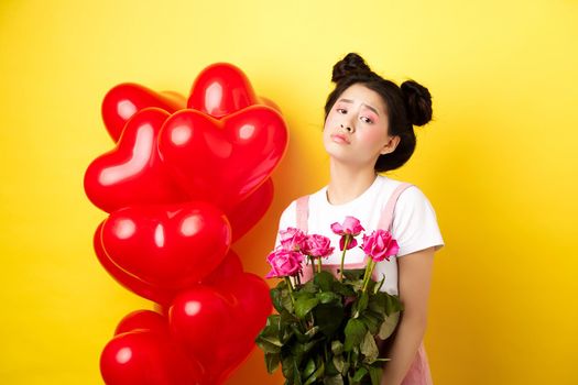 Happy Valentines concept. Sad and gloomy asian woman holding bouquet of roses and feeling upset and lonely on romantic lovers day, standing near red heart balloons, yellow background.