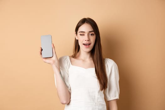 Cheerful stylish girl showing smartphone app, demonstrate empty phone screen and winking, standing on beige background.