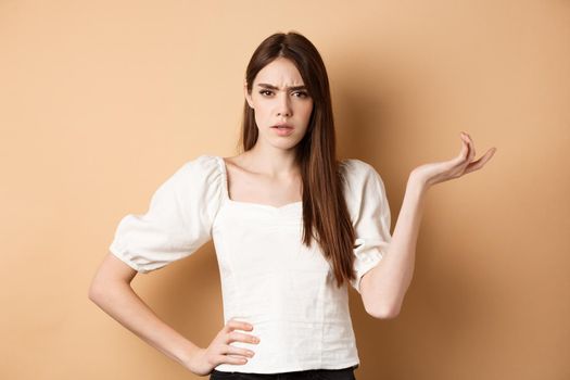 So what. Confused and annoyed young woman raising hand up and staring bothered at camera, cant understand wtf going on, standing on beige background.