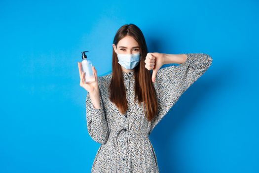 Covid-19, social distancing and healthcare concept. Disappointed girl in medical mask showing bad hand sanitizer and thumbs down, standing on blue background.