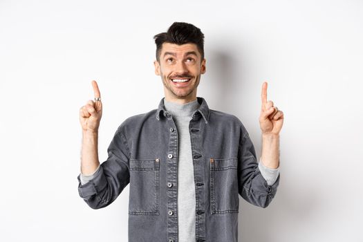 Cheerful caucaisan man with moustache pointing and looking up, smiling dreamy, showing cool promotion deal, standing on white background.