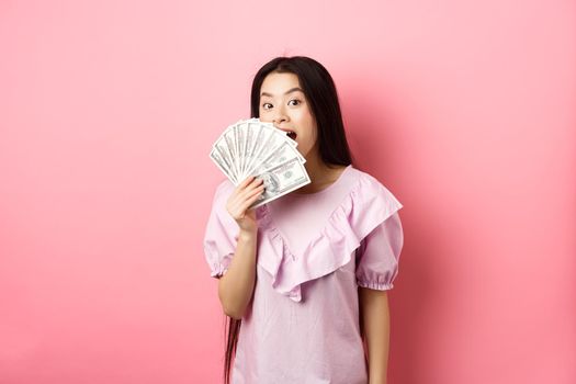 Cute asian woman showing dollar bills and smiling amazed. Rich woman waiving with money, standing against pink background.