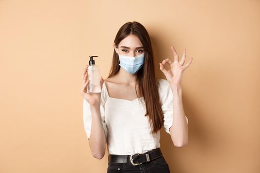 Covid-19 and preventive measures concept. Beautiful girl in face mask showing hand sanitizer bottle and okay sign, standing on beige background.