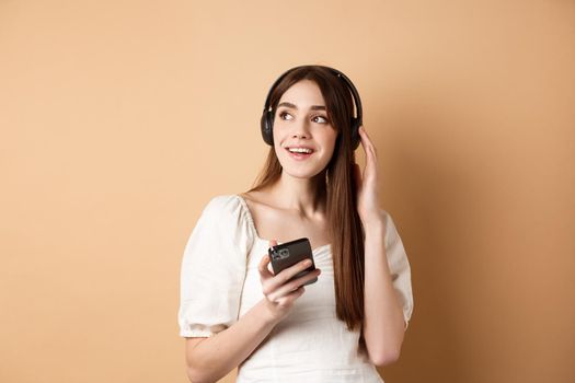 Attractive girl listening music in wireless headphones, holding mobile phone and looking aside with dreamy smile, beige background.