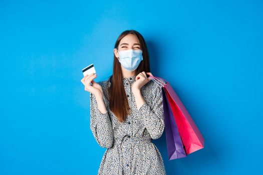 Covid-19, pandemic and lifestyle concept. Happy girl wear face mask on shopping, showing plastic credit card and laughing carefree, buying gifts, standing on blue background.