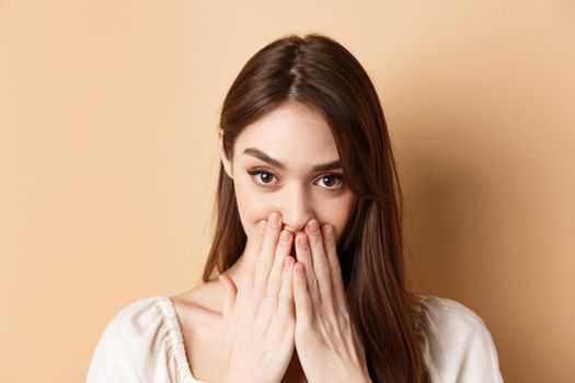 Close up of coquettish pretty girl covering mouth, looking happy and romantic, standing on beige background.