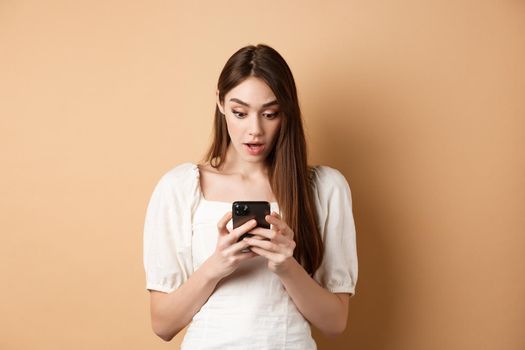 Woman look surprised at smartphone screen, reading shocking message on cell phone, standing amazed on beige background.