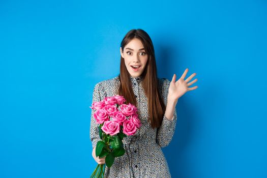 Valentines day concept. Surprised pretty girl holding bouquet of flowers from lover, looking amazed and happy at camera, standing on blue background.