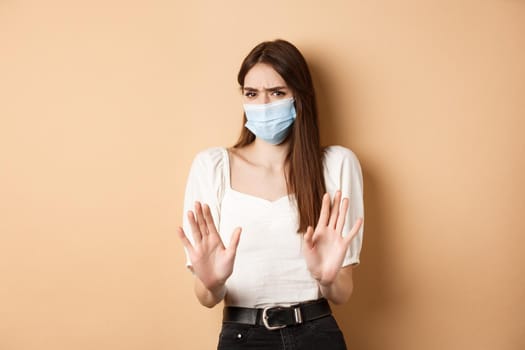 Pandemic and healthcare concept. Keep distance. Young woman in medical mask stretch out hand in stop sign, telling to stay away and frowning disgusted, beige background.