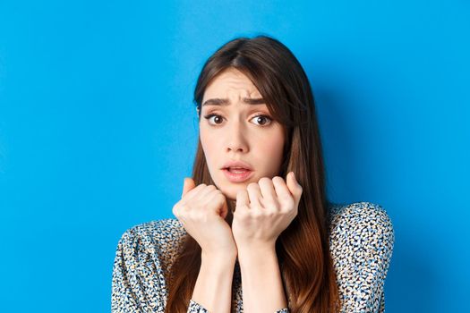 Close-up of scared timid girl looking afraid, shaking from fear, press hands to body and staring frightened, standing on blue background.