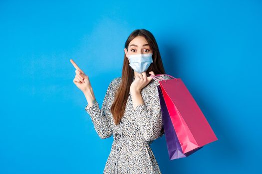 Covid-19, pandemic and lifestyle concept. Excited woman wears medical mask on shopping, pointing left corner logo, holding bags over shoulder, blue background.