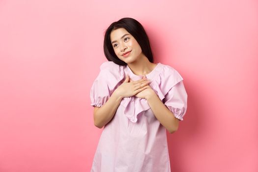 Dreamy asian girl smiling and holding hands on chest, looking left at something romantic and cute, standing on pink background.