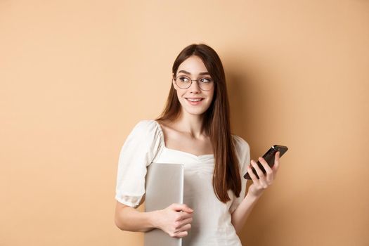 Image of young stylish woman going on work, holding laptop and smartphone, looking aside at empty space, standing on beige background.